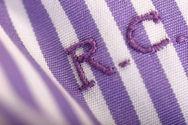 Close-up of a purple and white striped shirt with 'R.C.' initials embroidered in purple thread on the left chest, highlighting detailed stitching.