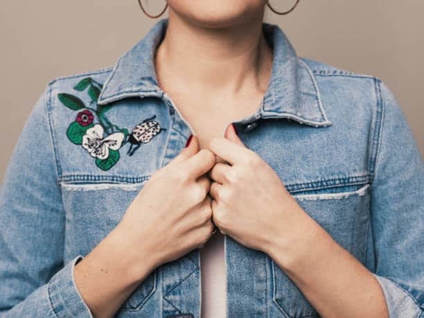 Person wearing a denim jacket with floral embroidery on the left chest, featuring white flowers and a ladybug design, with hands clasped in front.