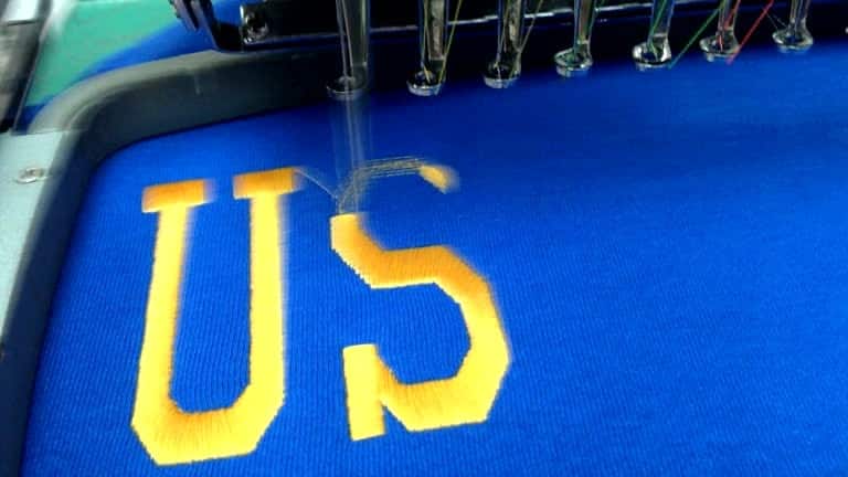 Embroidery machine stitching the letters 'US' in yellow thread on blue fabric, demonstrating the process of creating a left chest logo with precise sizing.