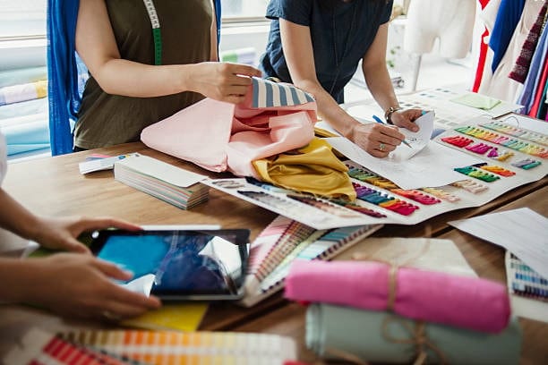 Designers selecting from various cloth fabric types like silk and cotton, working with swatches and rolls in an apparel studio.