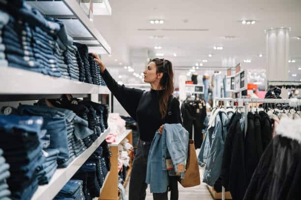 Woman browsing stacks of jeans in a store, considering fabric yardage needs for pants sewing projects.