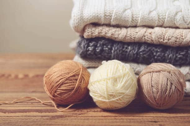 Balls of wool yarn in front of stacked sweaters, representing a natural fiber cloth fabric type for cozy apparel.