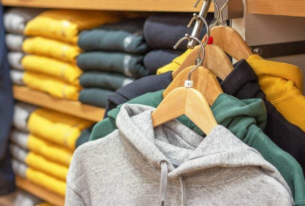 Hoodies in various colors displayed on hangers with folded sweatshirts in the background, comparing hoodie and sweatshirt options