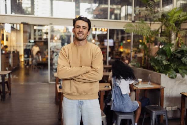 Smiling man in a beige sweatshirt standing inside a café, illustrating casual and versatile sweatshirt styling
