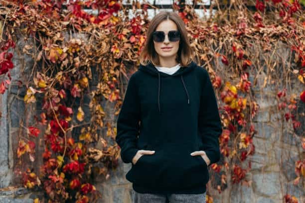 Woman in a black hoodie and sunglasses standing against autumn leaves, showcasing cozy and stylish hoodie fashion